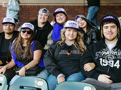 A group of seven people smiling while seated at Oracle Park wearing co-branded SFSU/San Francisco Giants baseball caps