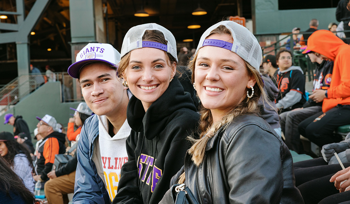 Three people smile while seated and wearing co-branded SFSU-San Francisco Giants baseball caps at Oracle Park