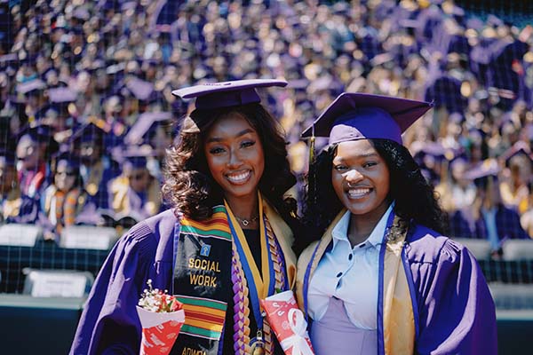 Two graduates smile and pose for a picture in graduation regalia in front of the crowd at SFSU Commencement
