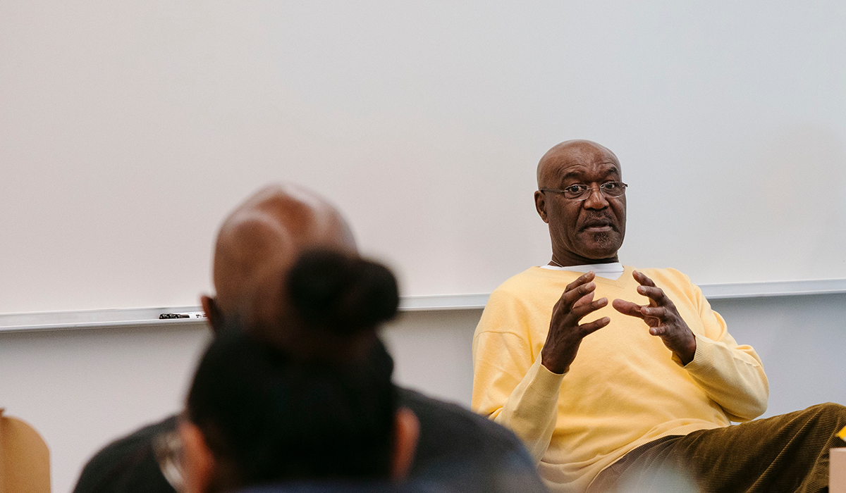 Delroy Lindo cups his hands together while seated in front of a whiteboard in a classroom wearing eyeglasses and a yellow sweater