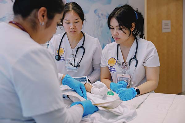 Three students in San Francisco State University scrubs, stethoscopes and rubber gloves handle a doll of a newborn baby
