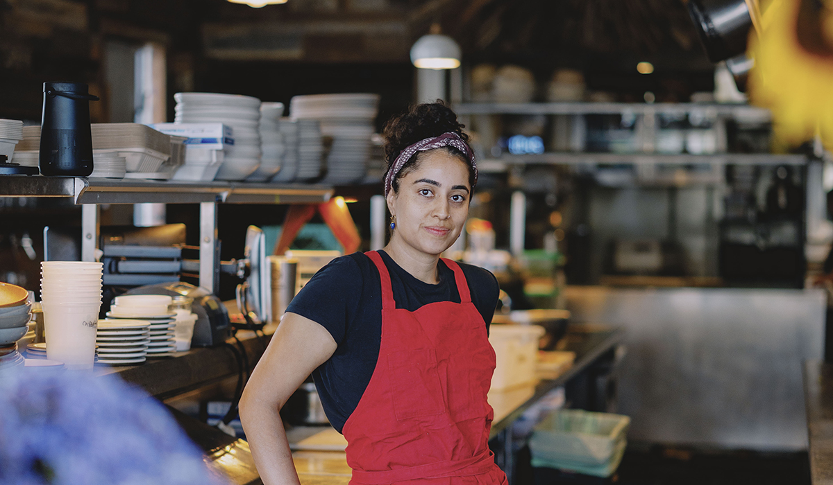 A Person in a red apron poses for a photo while standing inside a commercial kitchen