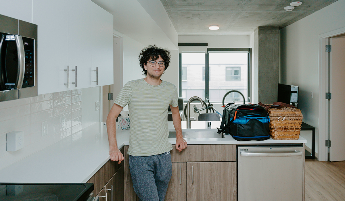 A student leans against the counter in his kitchenette in a residence hall with bags, a bicycle and a window behind him