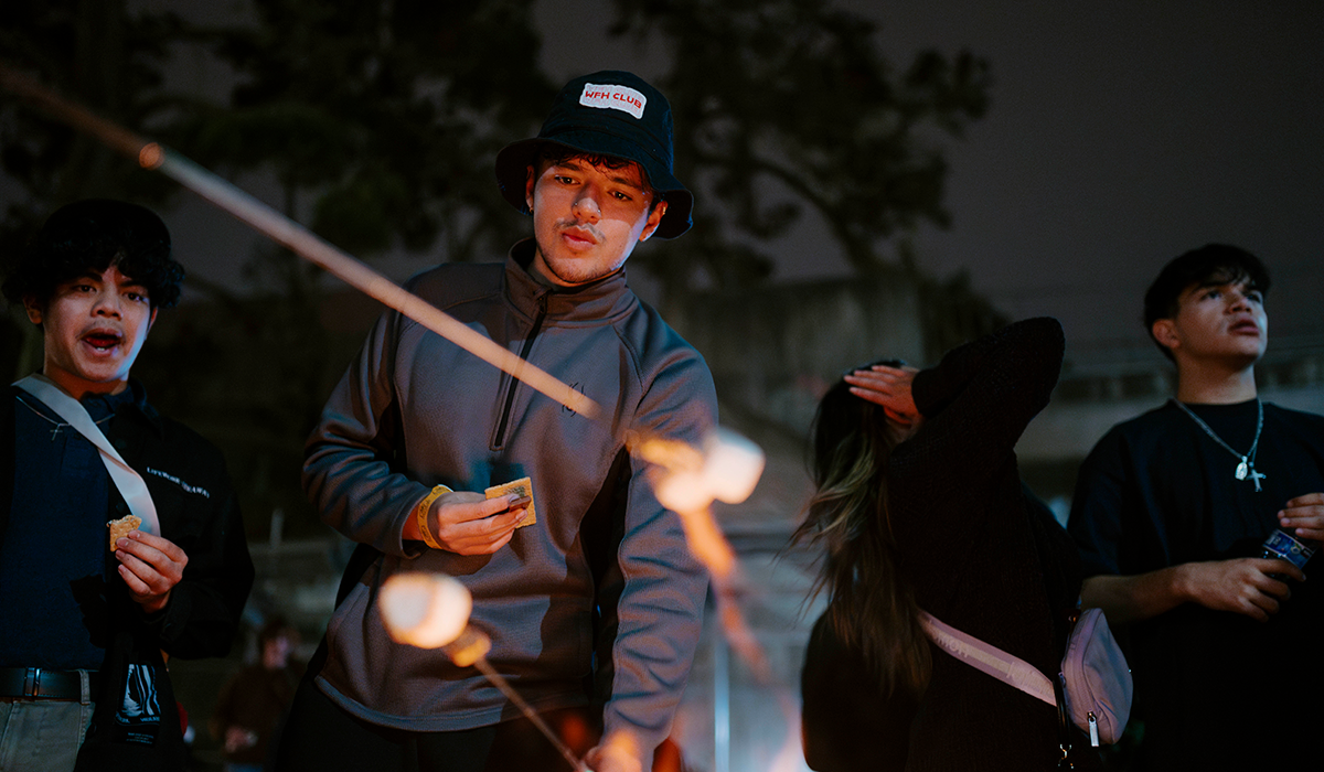 At an evening bonfire, a person lights a marshmallow on a stick while others watch and eat S'Mores