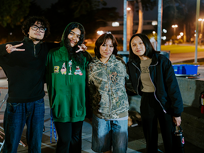 Four students pose for a photo while standing outdoors at night with one of them flashing a peace sign