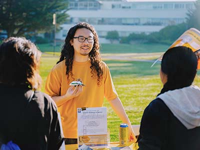 A student in a yellow shirt stands behind a table talking to students and holding a partially eaten Clif Bar