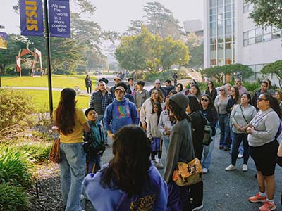 A student tour guide speaks to a group of families on a campus tour while standing on a curb near Burk Hall