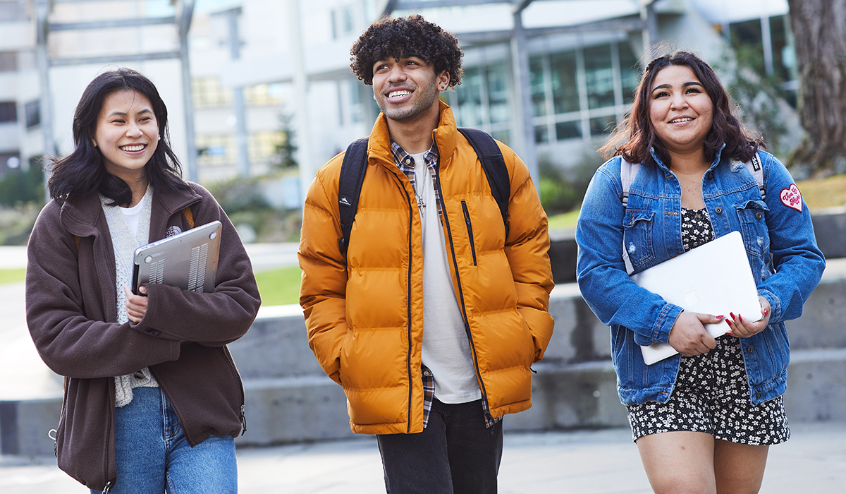 Three students walking in a line on a university campus while they are smiling.