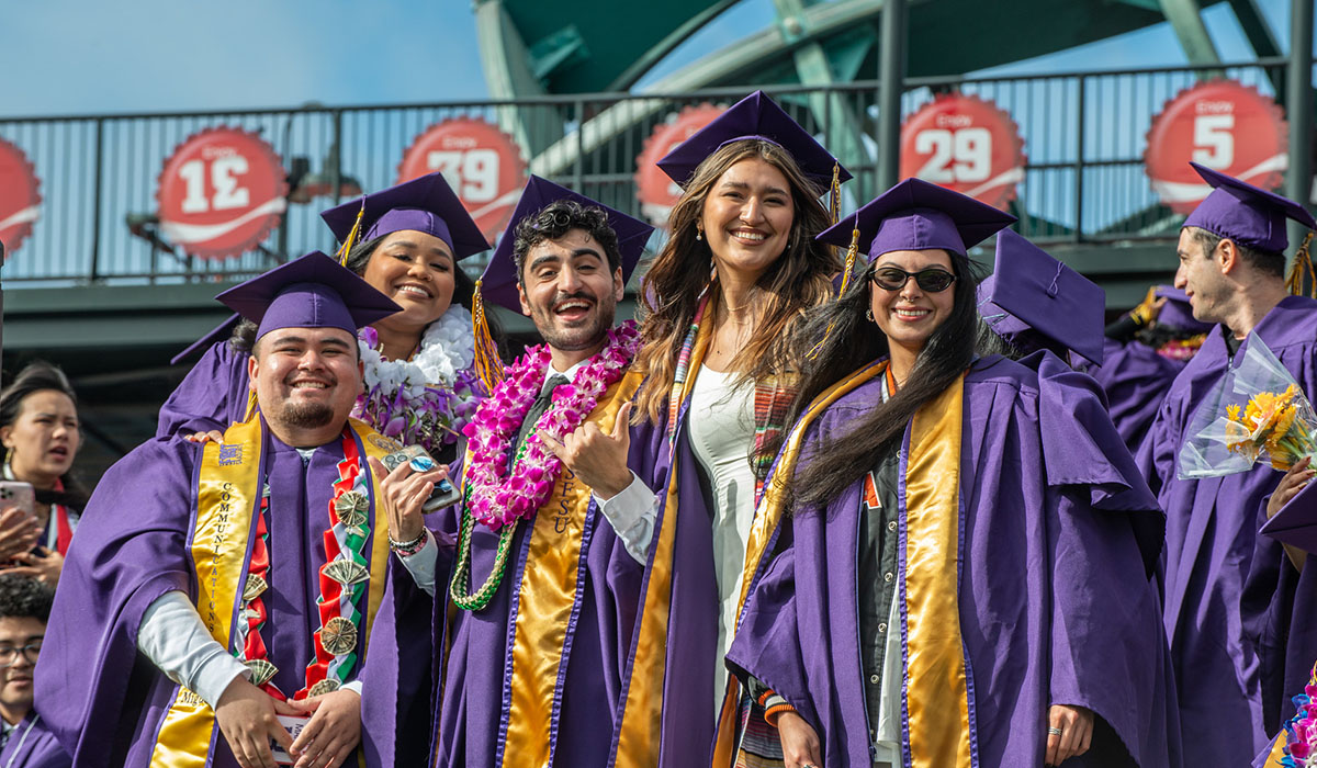 Graduates dressed in regalia stand together in the bleachers at Oracle Park