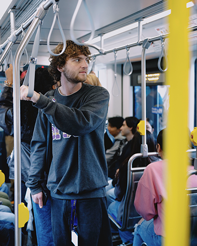 A student wearing an SFSU sweatshirt and a backpack holds a pole while standing on a Muni light-rail train surrounded by other passengers standing and sitting
