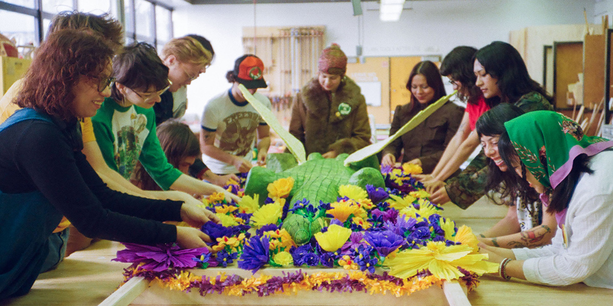 About 12 students in an art studio work on a sculpture of an alligator surrounded by flowers and laying on a stretcher