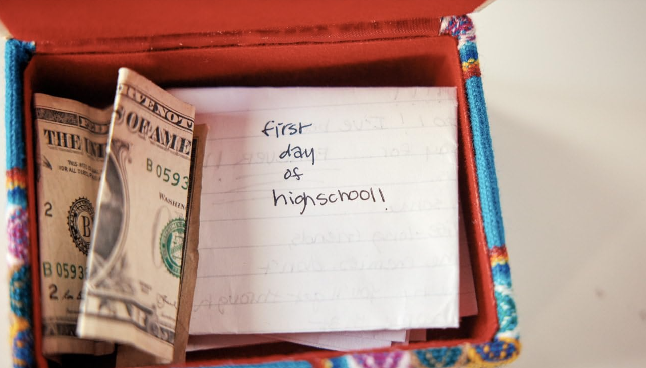 A box contains a dollar bill and a handwritten note saying "first day of high school"