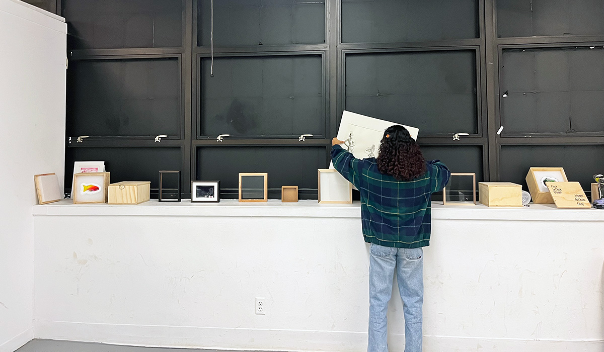 A student is standing in a room, facing away from the camera, organizing or examining items on a counter beneath large wall-mounted cabinets. The cabinets are open, displaying empty shelves. Atop the counter are various books and small boxed items. 