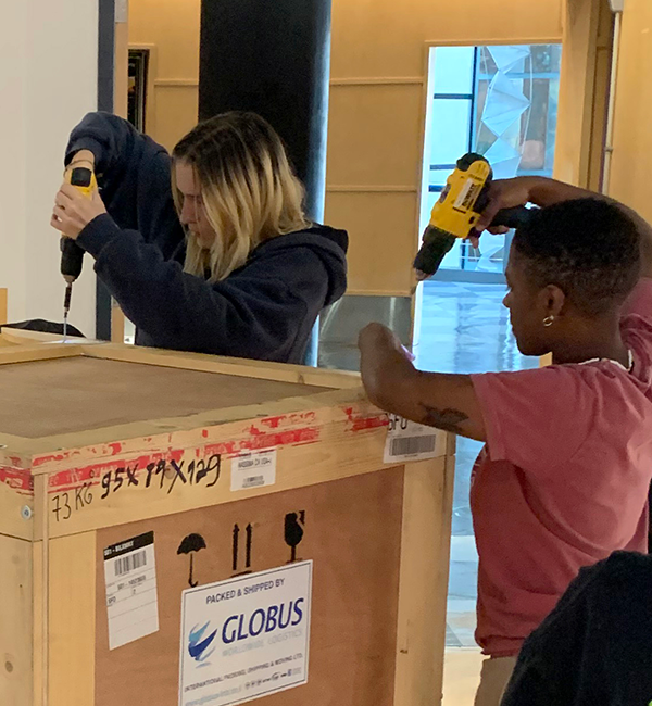 Two students drill into a 5-foot-tall wooden box while standing in the Fine Arts Gallery