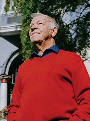 Bob Buckter smiles while standing in front of a Victorian home and a tree in San Francisco while wearing a red sweater with a collared blue shirt underneath it