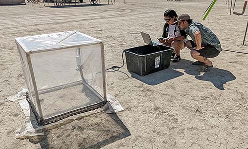 Two people on the desert floor looking at a computer near a large transparent cube