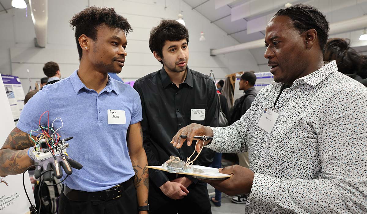 Two male students showing a robotic arm to a male judge