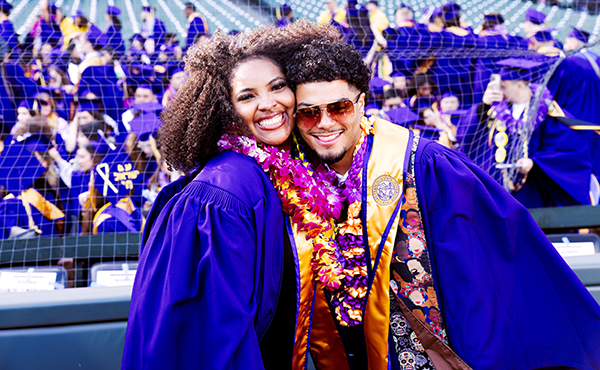 Two graduating students embrace while posing for a picture on the field at Oracle Park while other graduating students are in the stands behind them