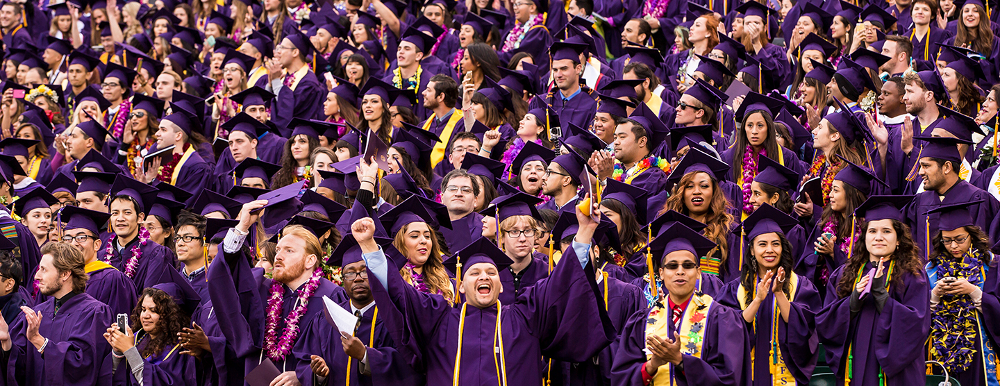 Several students stand in a stadium dressed in their caps and gown while one student smiles directly at the camera gesturing triumphantly