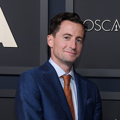 A person with short brown hair dressed in a blue suit with a red tie smiles.