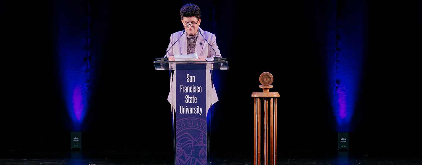 President Lynn Mahoney speaks behind a lectern decorated in purple with the text San Francisco State University in front of a black background and next to a wooden gavel 