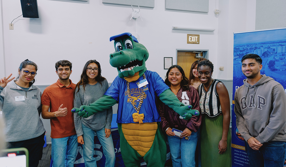 Seven people smile while posing for a photo with the Alli Gator mascot in the middle pointing their fingers horizontally and with one of the other people flashing a peace sign