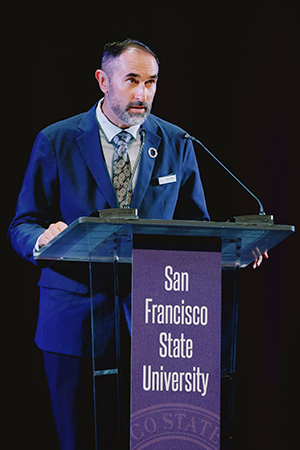 Jackson Wilson speaks from a San Francisco State University lectern while wearing a suit and tie