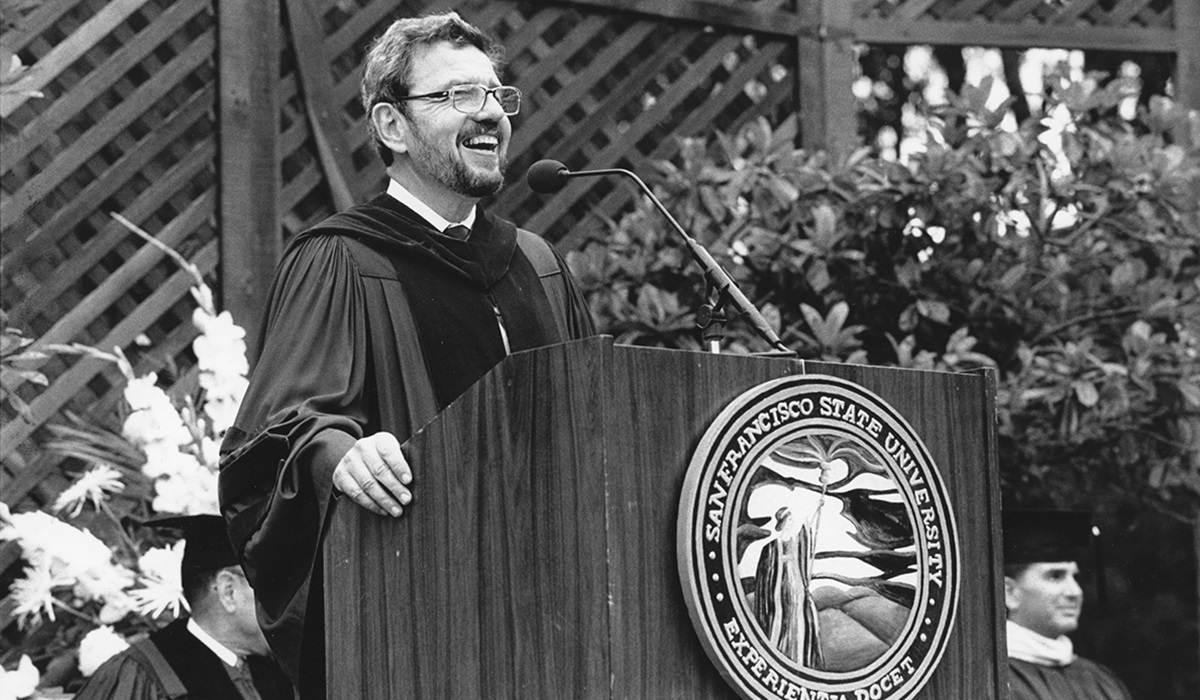 A smiling Robert Corrigan in Commencement robe standing at a podium