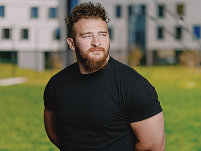 Julian Dancel stands in front of new student housing West Grove Commons with his arms folded behind his back and while wearing a black short-sleeve T-shirt
