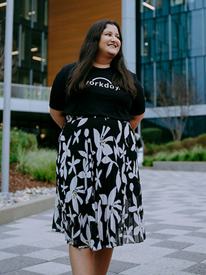 Shriya Dandin smiles and looks over her left shoulder while standing outside of an office building while wearing a Workday T-shirt and a skirt with floral designs