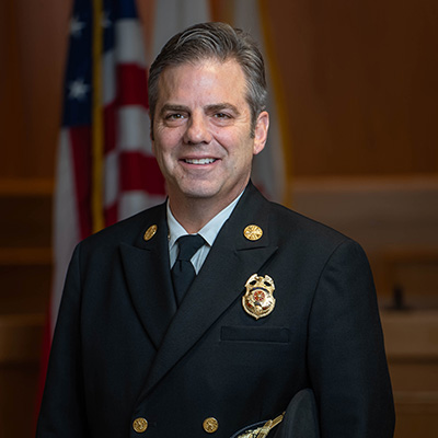 A person with short gray hair dressed in formal uniform poses for a photo with the American flag in the bacckground