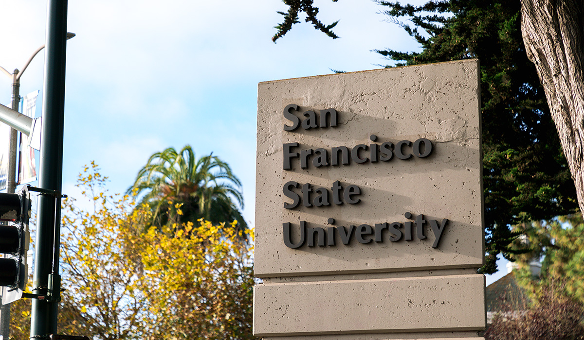 The San Francisco State University pylon sign surrounded by trees and next to a stoplight and a bicyclist road sign on a mostly sunny day