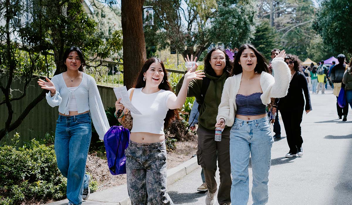 Four women waving to camera while walking on campus