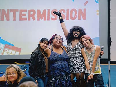 Three students smile and stand with a drag queen raising their right hand in the air