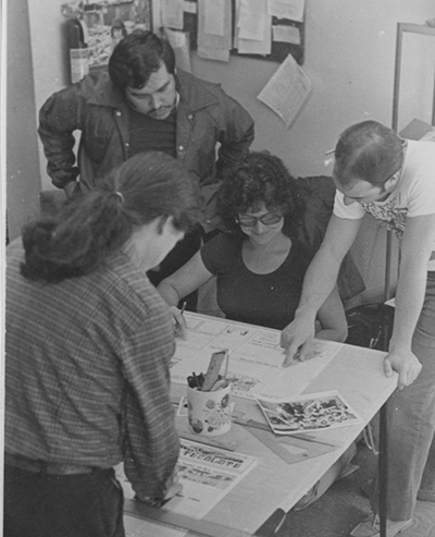 Juan Ginzales and other people crowd around a table to look at a newspaper layout