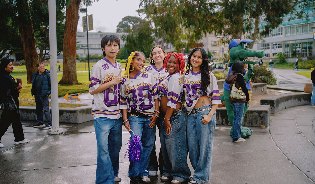 Five people wearing white and purple football jerseys and blue jeans stand on Malcolm X Plaza on an overcast and rainy day