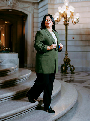 Kimberly Gallardo looks over her left shoulder while walking down a stairway in San Francisco City Hall and wearing an olive green sportscoat, a white shirt and black slacks