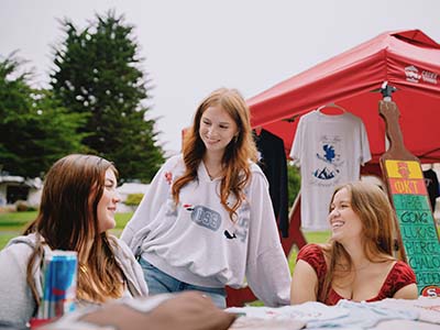 Three students talk at a table at the Gatorfest on the Quad next to a canopy with a T-shirt hanging from it