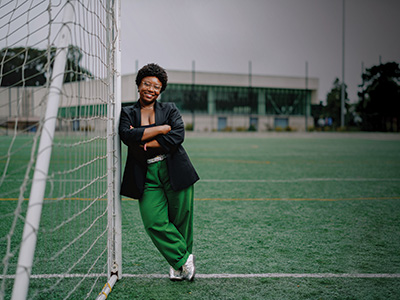 Tamerra Griffin leans on a soccer goal post with the Mashouf Wellness Center visible behind her on an overcast day