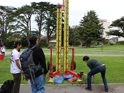 A person swings a hammer toward the ground while playing a hammer swing carnival game while two other people watch on the Quad on an overcast day