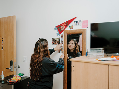 An SFSU student looks in the mirror while decorating the wall of her residence hall room with a pennant and pictures