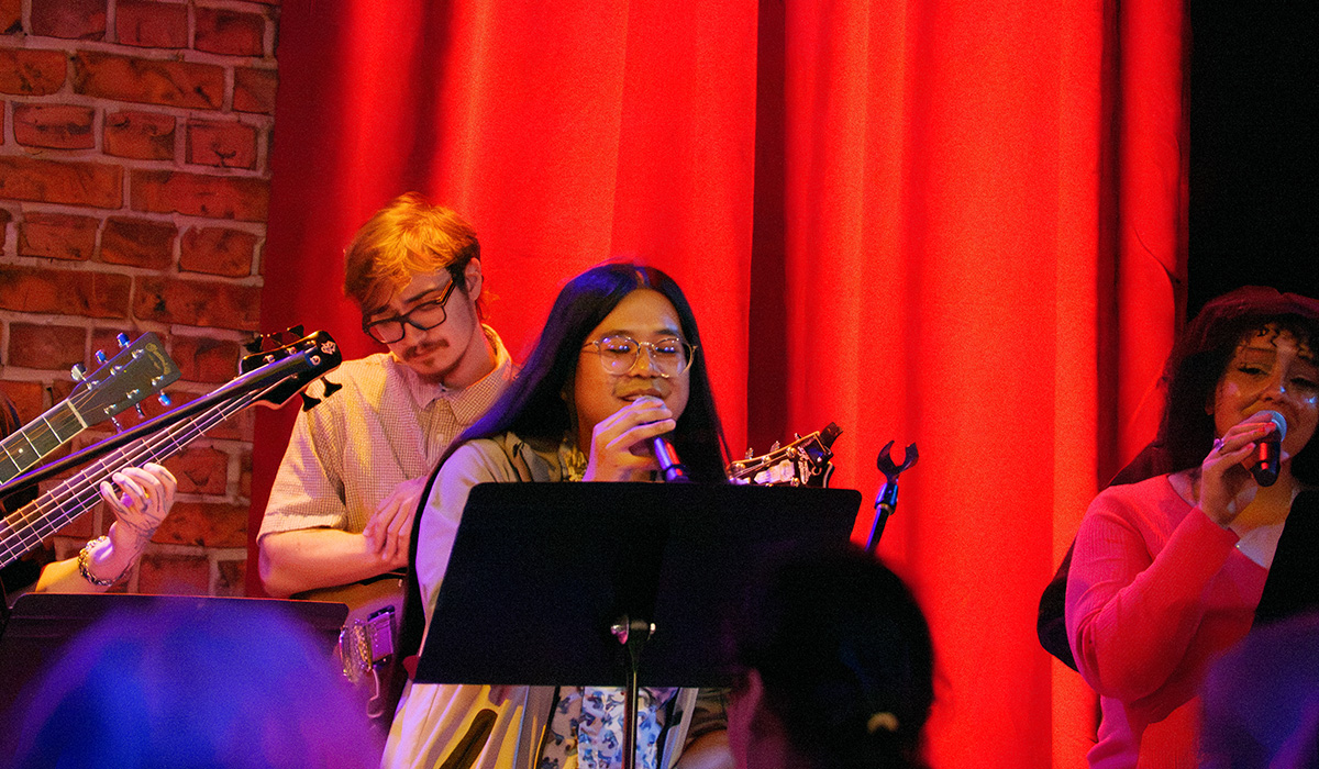 Anthony Joerel Garcia and Sarah Star sing while holding microphones and standing behind easels and in front of an electric guitarist and a red curtain