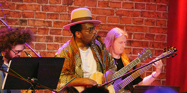 Martin Luther McCoy plays acoustic guitar while two students perform with him in front of a curtain decorated like a brick wall
