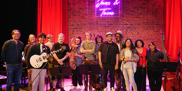 Jazz and Tacos performers and SFSU faculty members pose for a group photo in front of a curtain decorated to look like a brick wall and a neon sign with the cursive text Jazz and Tacos