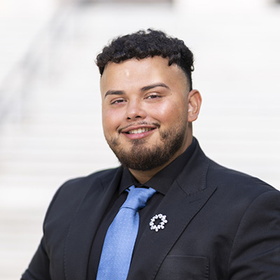 A person with short curl black hair and facial hair wearing a suit smiles. 