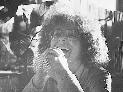 A black and white photo of Betty Kaplowitz giving a large smile while seated with her hands clasped, wearing eyeglasses and sitting in front of plants