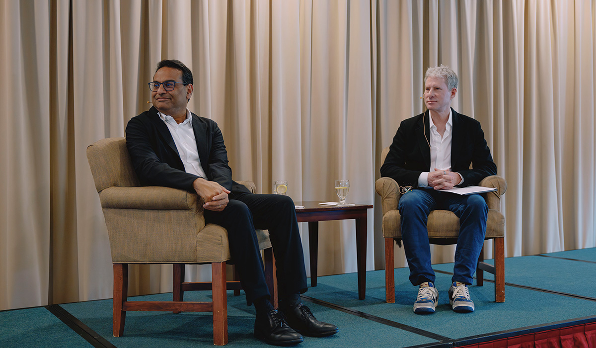 Laxman Narasimhan and Chris Larsen look to their right while seated on a stage between a table and in front of a curtain