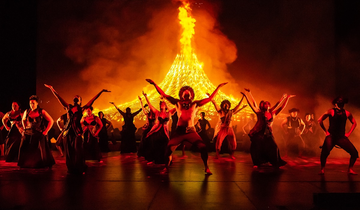 Dancers in traditional Hawaiian dress pose in front of a large fire