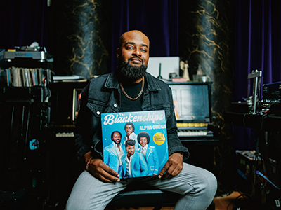 Mike Blankenship holds a vinyl record of The Blankenships while seated in front of a piano inside of a recording studio and wearing a black shirt, necklace, jean jacket and jean pants