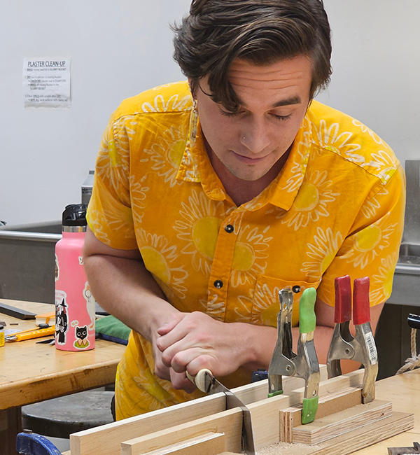 Adrian Morelock-Revon cuts into wooden blocks while standing and wearing a yellow short-sleeved collared shirt. A pink reusable water bottle decorated with stickers and a whiteboard are visible behind Morelock-Revon